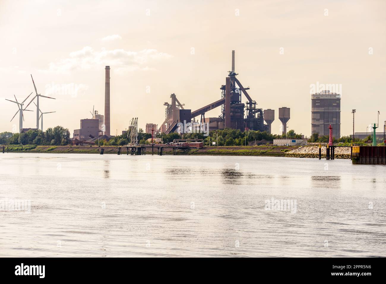 Riverside integrated steel plant at sunset in summer. Wind turbines are in background. Stock Photo