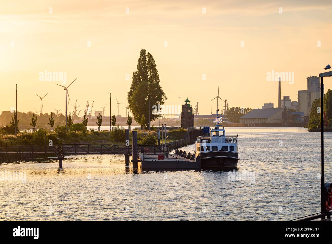River harbour at sunset in summer. Manufacturing plants and wind turbines along with a lighthouse are visible in background. Stock Photo