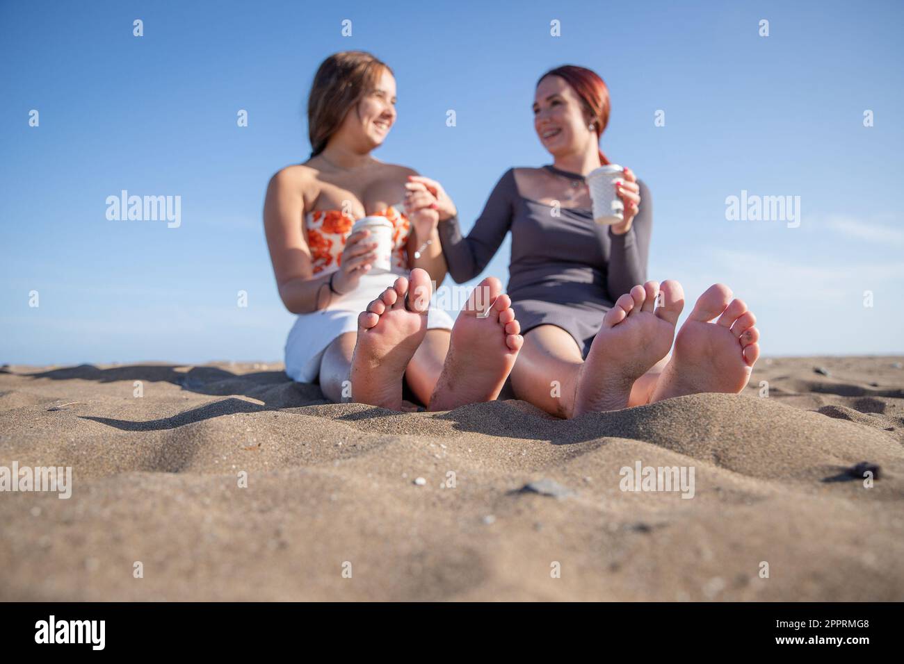 Two girlfriends drink coffee sitting at the beach, close-up on their feet Stock Photo