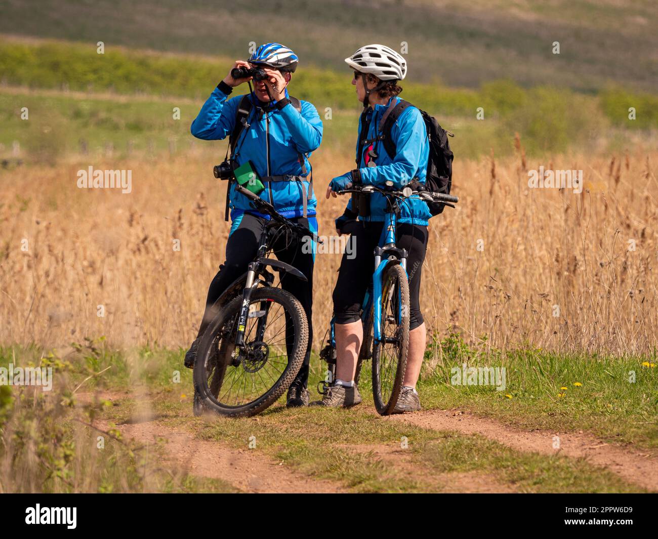 Two adult mountain bikers pausing on a grass track to look through binoculars at the surrounding countryside. St Aidan's Park. Leeds Stock Photo