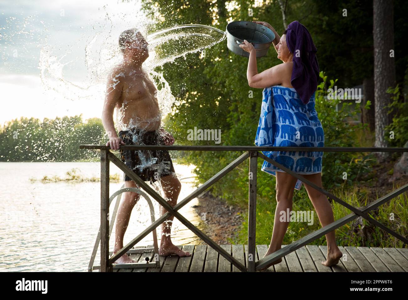 Aged couple having fun after Finnish sauna on wooden cottage pier in a lake. Mature woman pouring cold water from basin over her partner. Finland Stock Photo