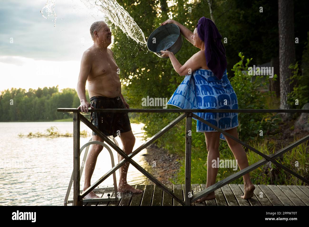 Aged couple having fun after Finnish sauna on wooden cottage pier in a lake. Mature woman pouring cold water from basin over her partner. Finland Stock Photo