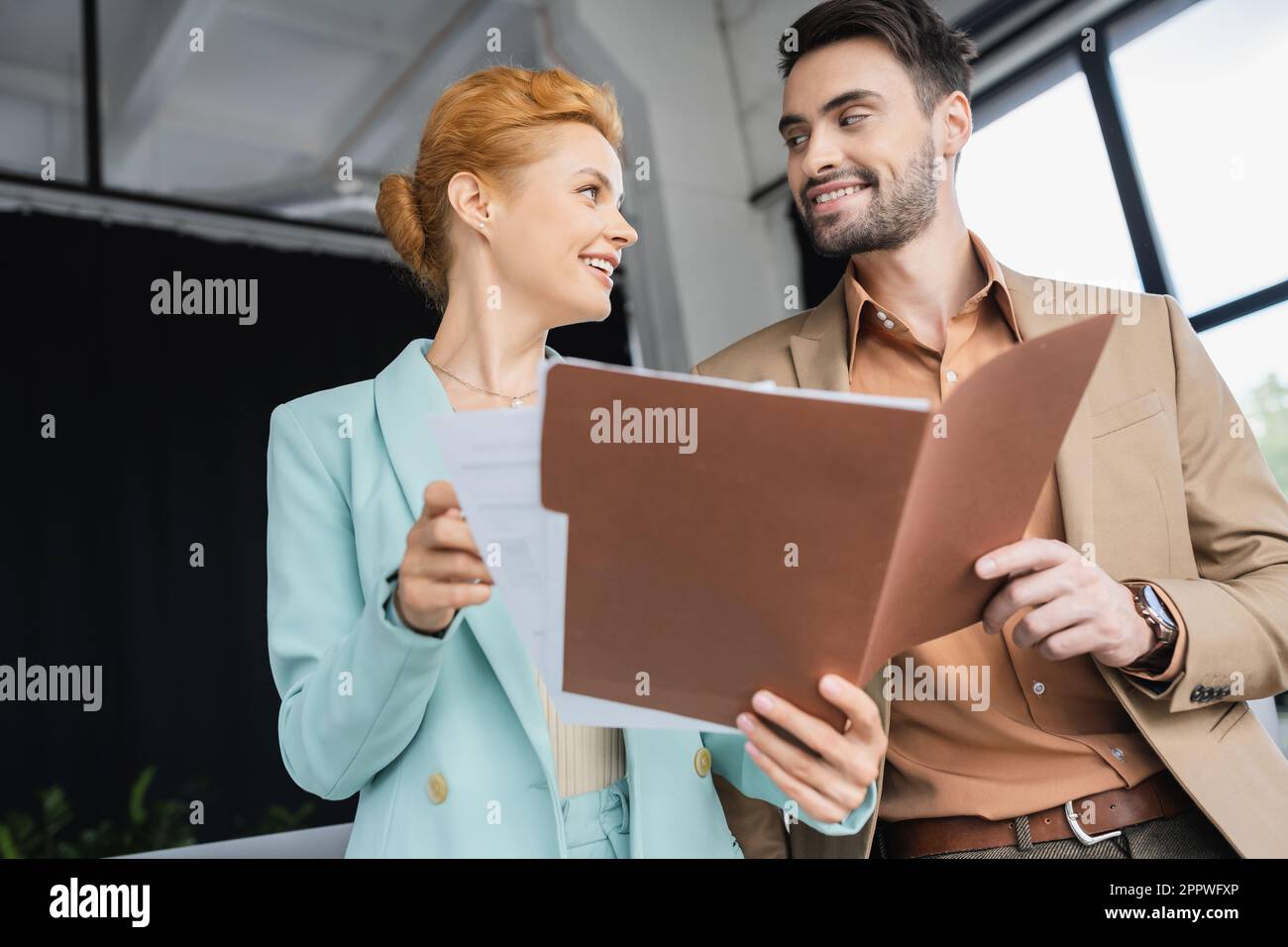 joyful business partners looking at each while talking near folder with papers in office,stock image Stock Photo