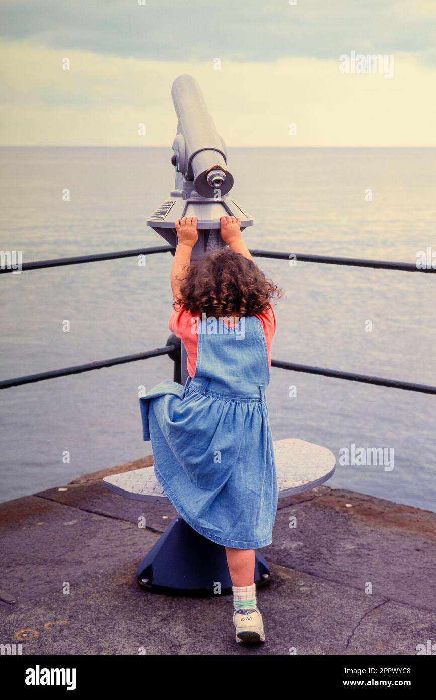 Rear view of a young caucasian child climbing up to the platform of a coin operated telescope at the seaside. Madeira. Stock Photo