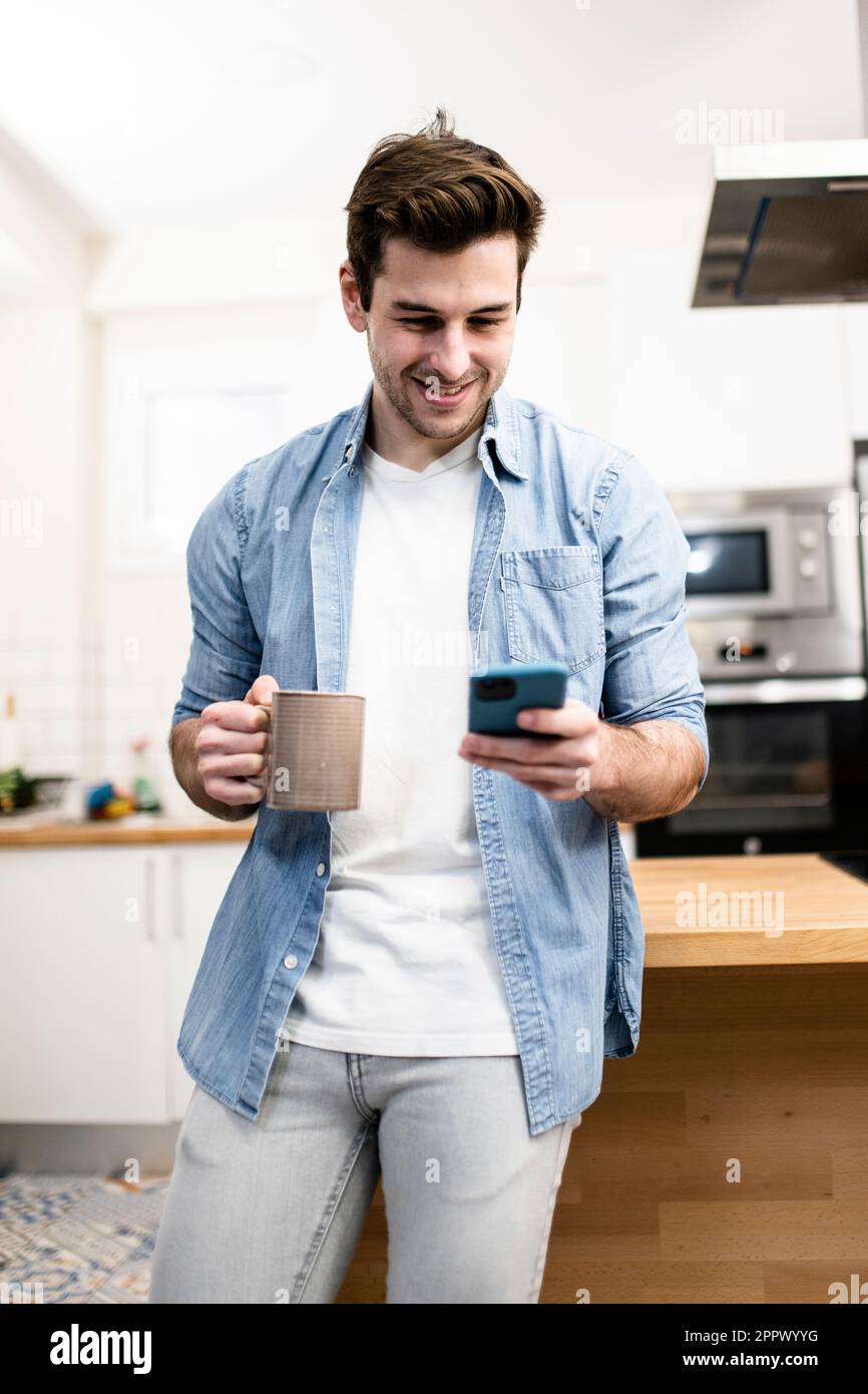 handsome happy man looking at his phone with a cup of tea in the kitchen. young man standing relaxed at home. Lifestyle concept. Stock Photo