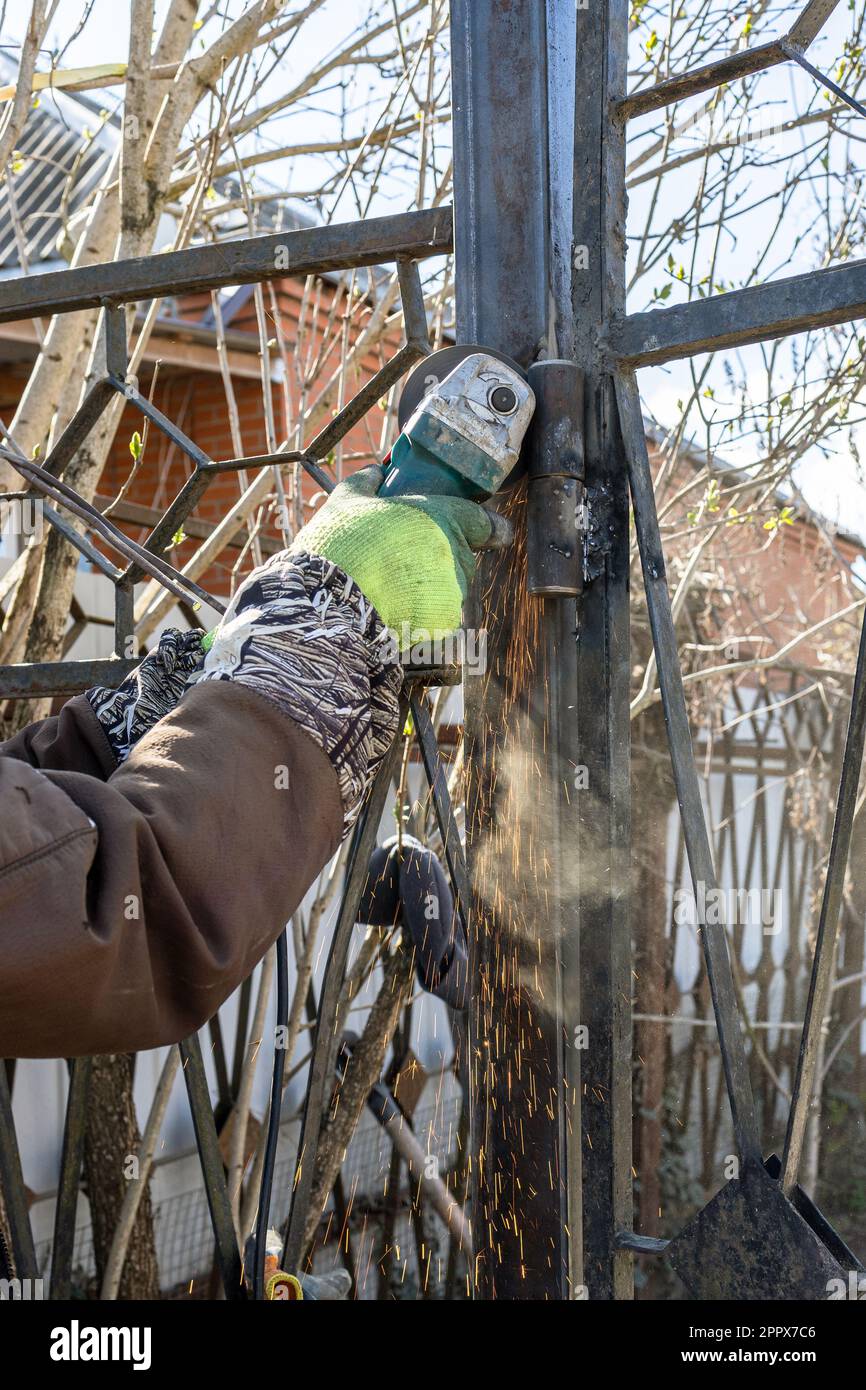 worker cutting hinge on gate of fence using electric grinder outdoors in village Stock Photo