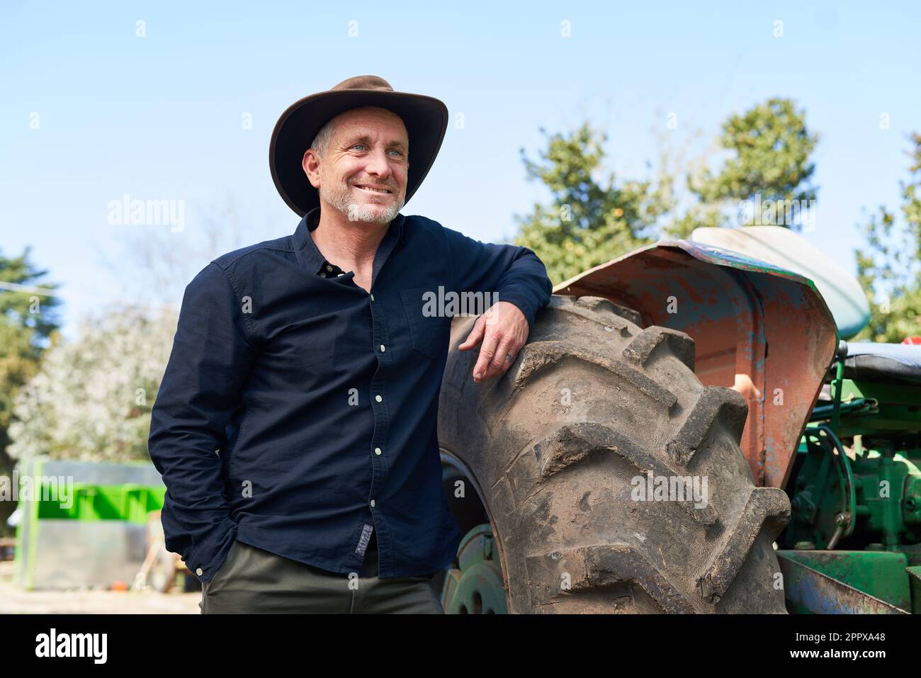 Happy mature male farmer leaning on tractor with clear sky in background during sunny day Stock Photo