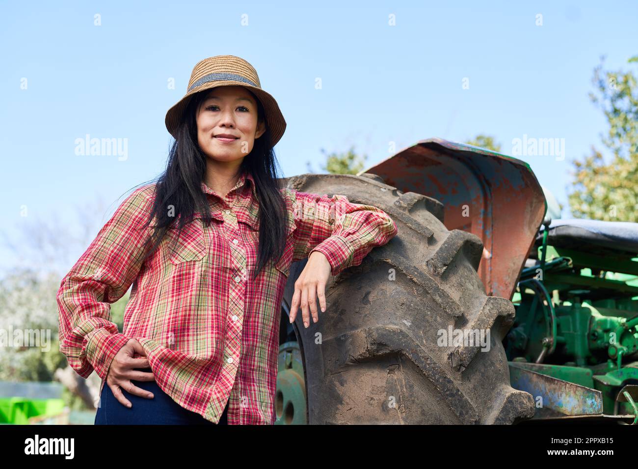 Portrait of smiling female farmer leaning on tractor with clear sky in background during sunny day Stock Photo