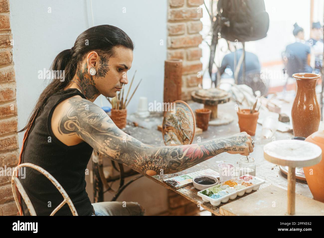 Concentrated young tattooed male artisan with long dark hair creating  picture on clay plate at table with various tools in workshop Stock Photo -  Alamy