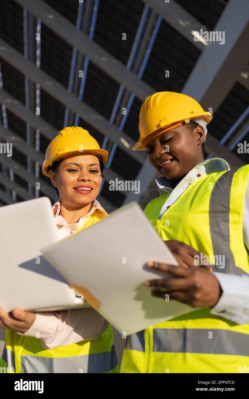 Low angle of young African American women in hardhats and engineer vests smiling and looking at documents while working on construction site Stock Photo