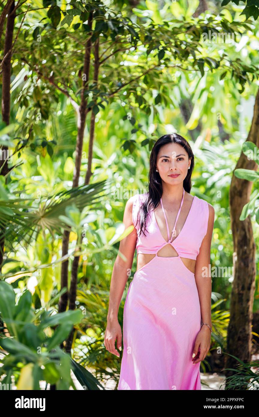 Calm young Mexican and Japanese female wearing pink summer maxi dress  looking at camera while standing in sunny green forest with palms and trees  in T Stock Photo - Alamy