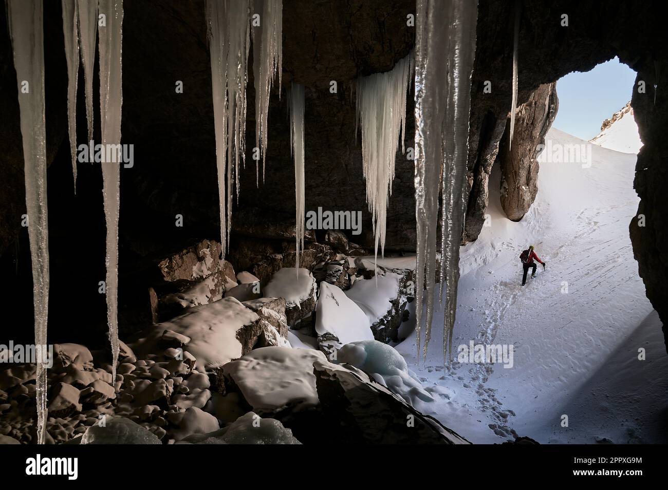 Back view of anonymous tourist with backpack and in warm clothes walking on snowy ground behind icicles while spending time alone at winter day in Gar Stock Photo