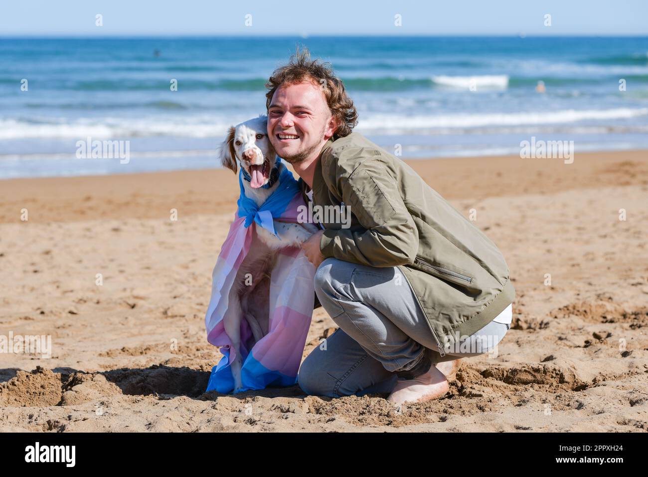 Friendly transgender man after transitioning embracing cute white Clumber Spaniel puppy wrapped in transgender pride flag relaxing on sandy beach on s Stock Photo