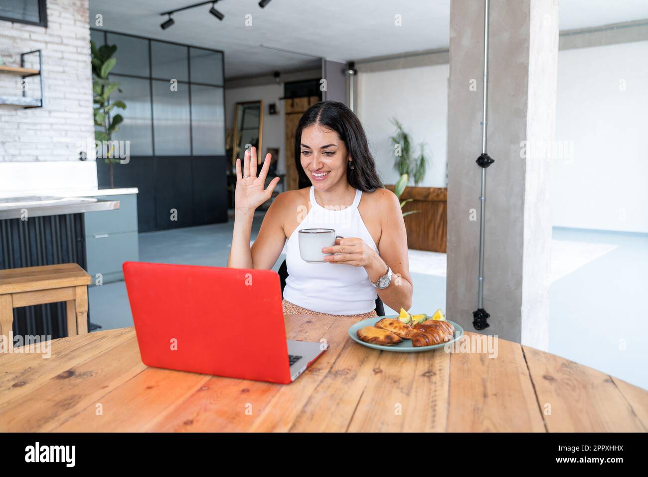 Young cheerful Hispanic female in white top sitting at wooden table with cup of hot drink croissant empanadas and avocado toasts while waving hand dur Stock Photo