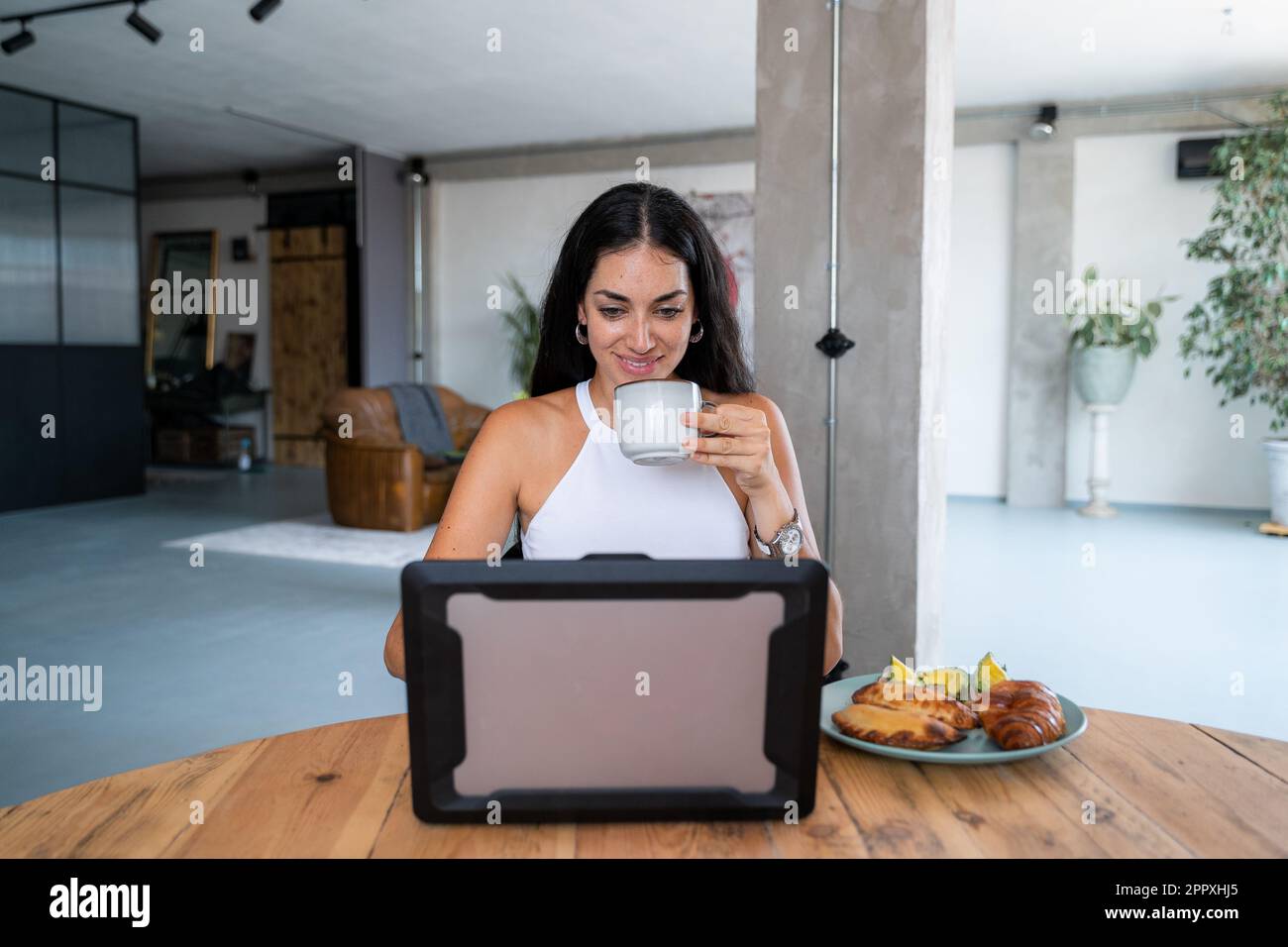 Young cheerful Hispanic female sitting at table drinking hot coffee having empanadas croissant and avocado toasts for breakfast while browsing netbook Stock Photo