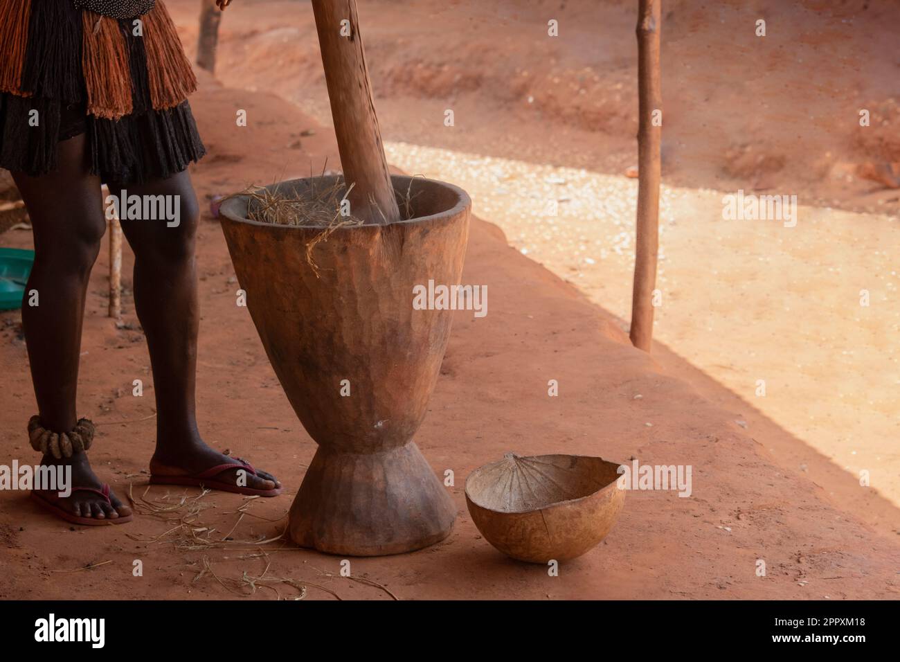 Crop anonymous African man in traditional clothes pestling plants in mortar during process of preparation of national meal on dusty street Stock Photo