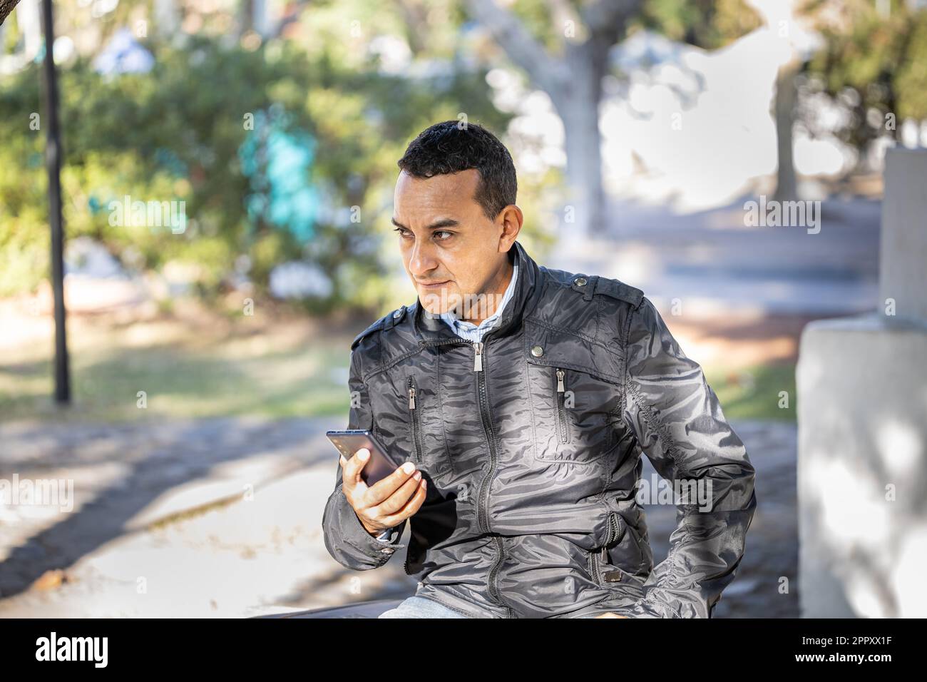 Young latin man sitting on a square bench using his mobile phone. Stock Photo