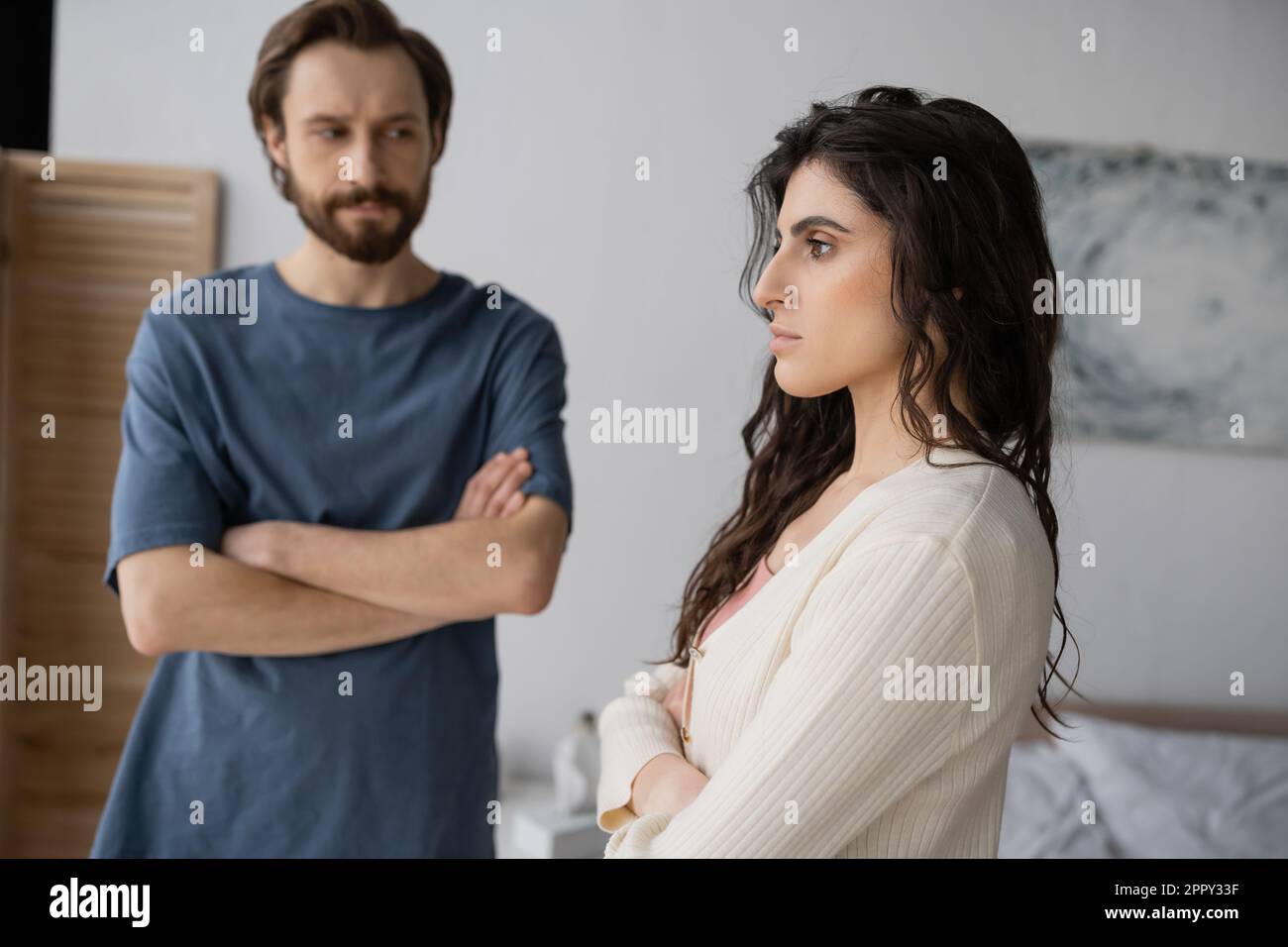 Offended brunette woman crossing arms near blurred boyfriend in bedroom,stock image Stock Photo