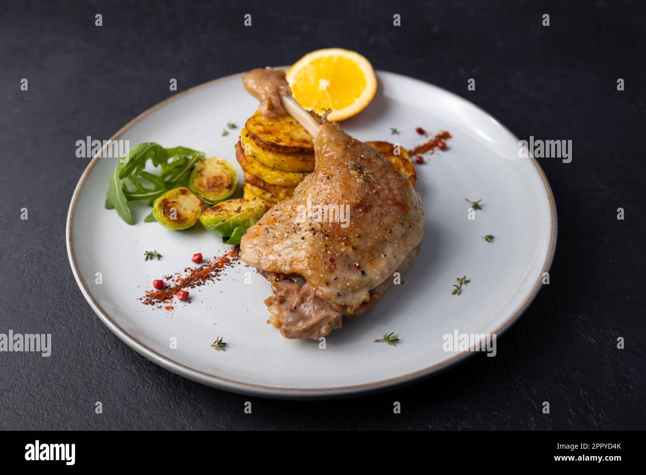 Duck leg confit with Brussels sprouts, baked potatoes, thyme and orange. Traditional French cuisine. Selective focus, close-up, black background. Stock Photo
