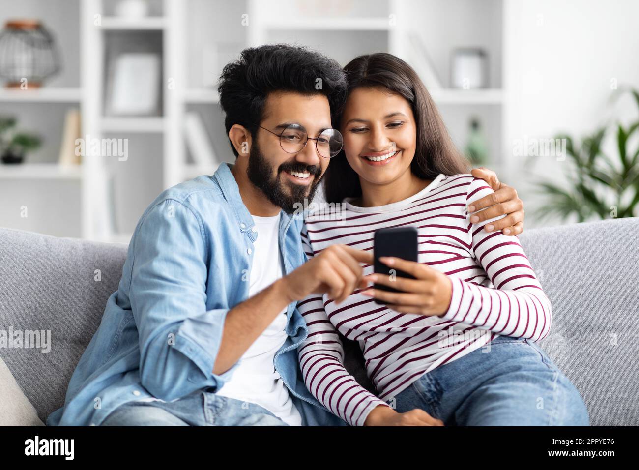 Relaxed multicultural couple sitting on sofa at home, using smartphone Stock Photo