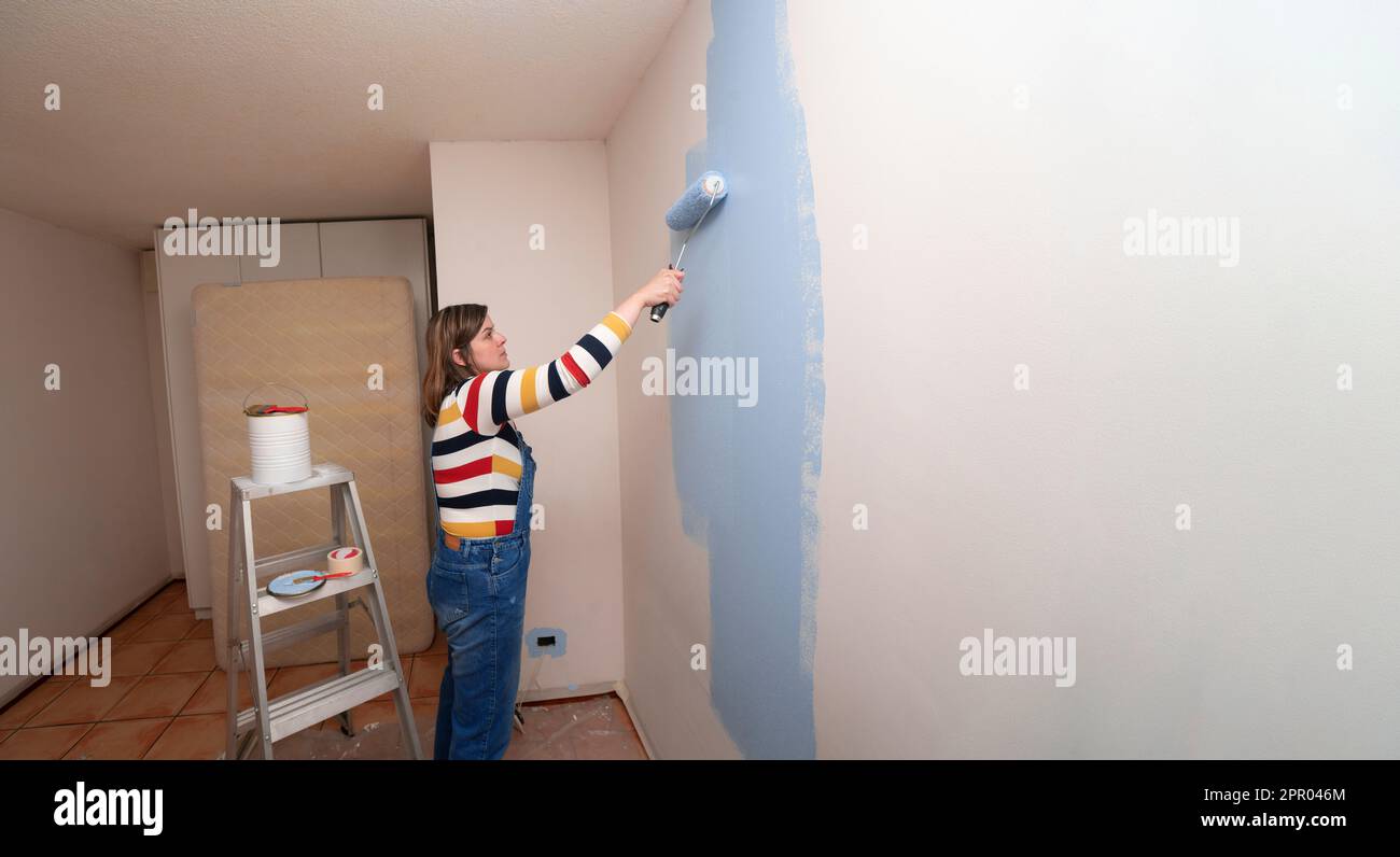 Standing beautiful woman dressed in overalls and striped blouse, profile view, with a roller in her hand painting a white wall with blue paint in an e Stock Photo