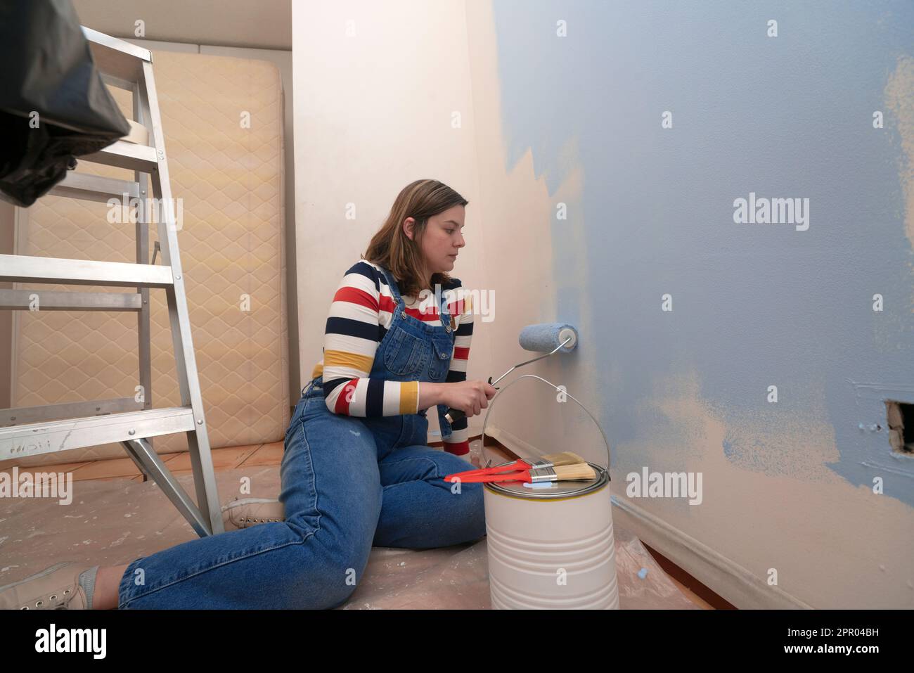 Kneeling woman dressed in overalls and striped blouse, profile view, painting a white wall with a roller with blue paint, inside an empty room with a Stock Photo