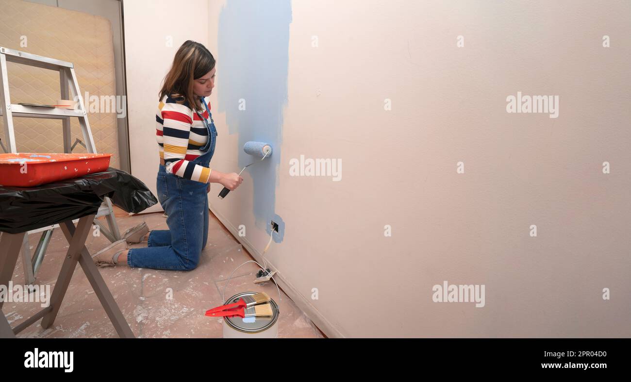 Kneeling woman dressed in overalls and striped blouse, profile view, painting a white wall with a roller with blue paint, inside an empty room with a Stock Photo