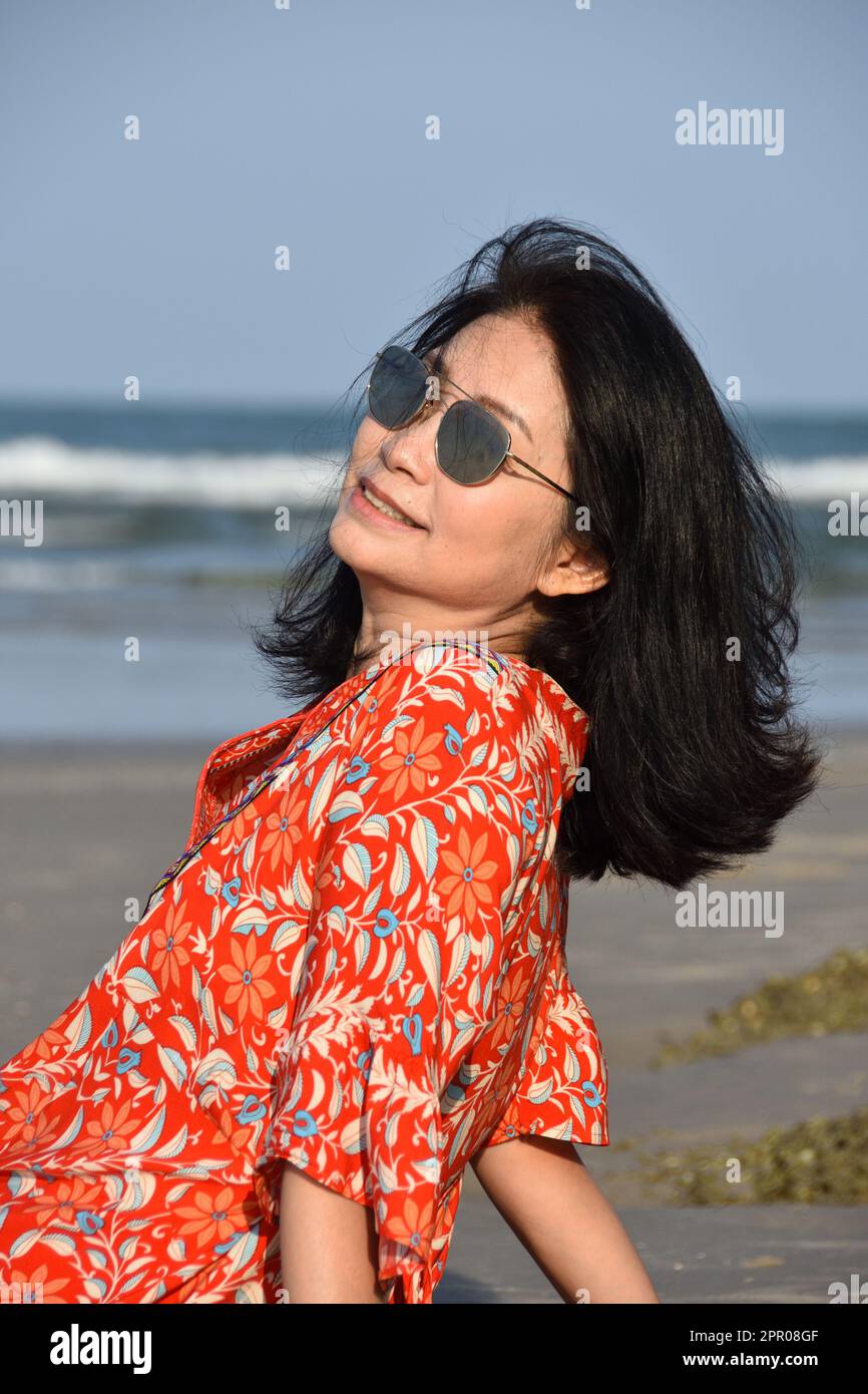 Ageingbetter2023 - A beautiful senior Asian woman sitting on the beach on a nice sunny day smiling and enjoying herself. Stock Photo