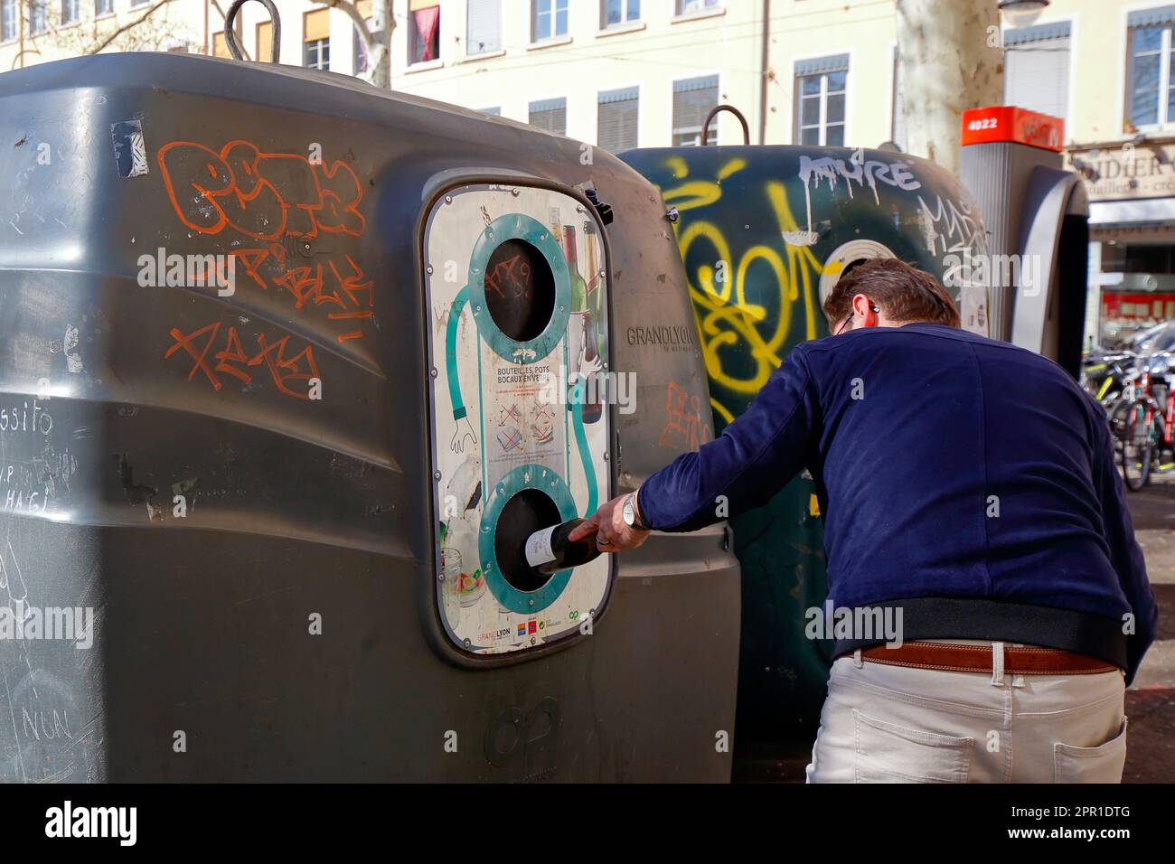 A person deposits an empty wine bottle into a large municipal recycling bin for glass bottles in