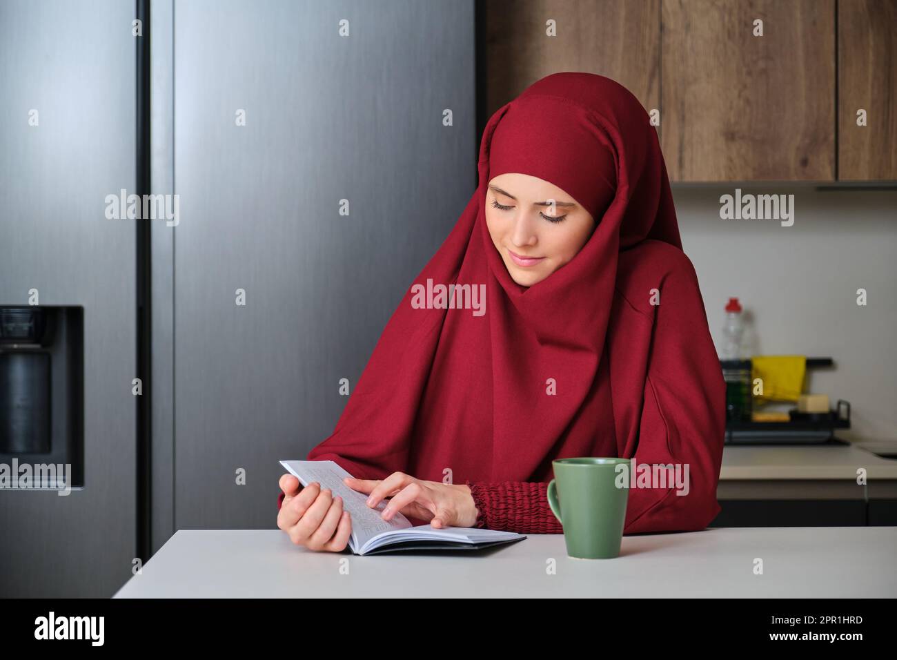 Muslim young woman in hijab drinking coffee and reading a book at home ...
