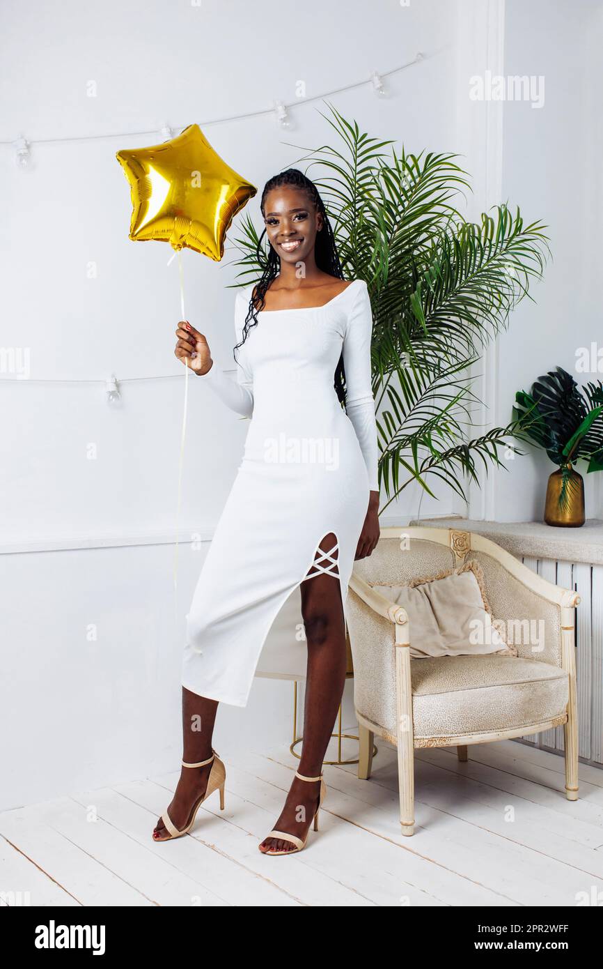 Smiling, elegant, shiny modern African American woman in white bodycon dress hold golden star balloon on holiday party Stock Photo