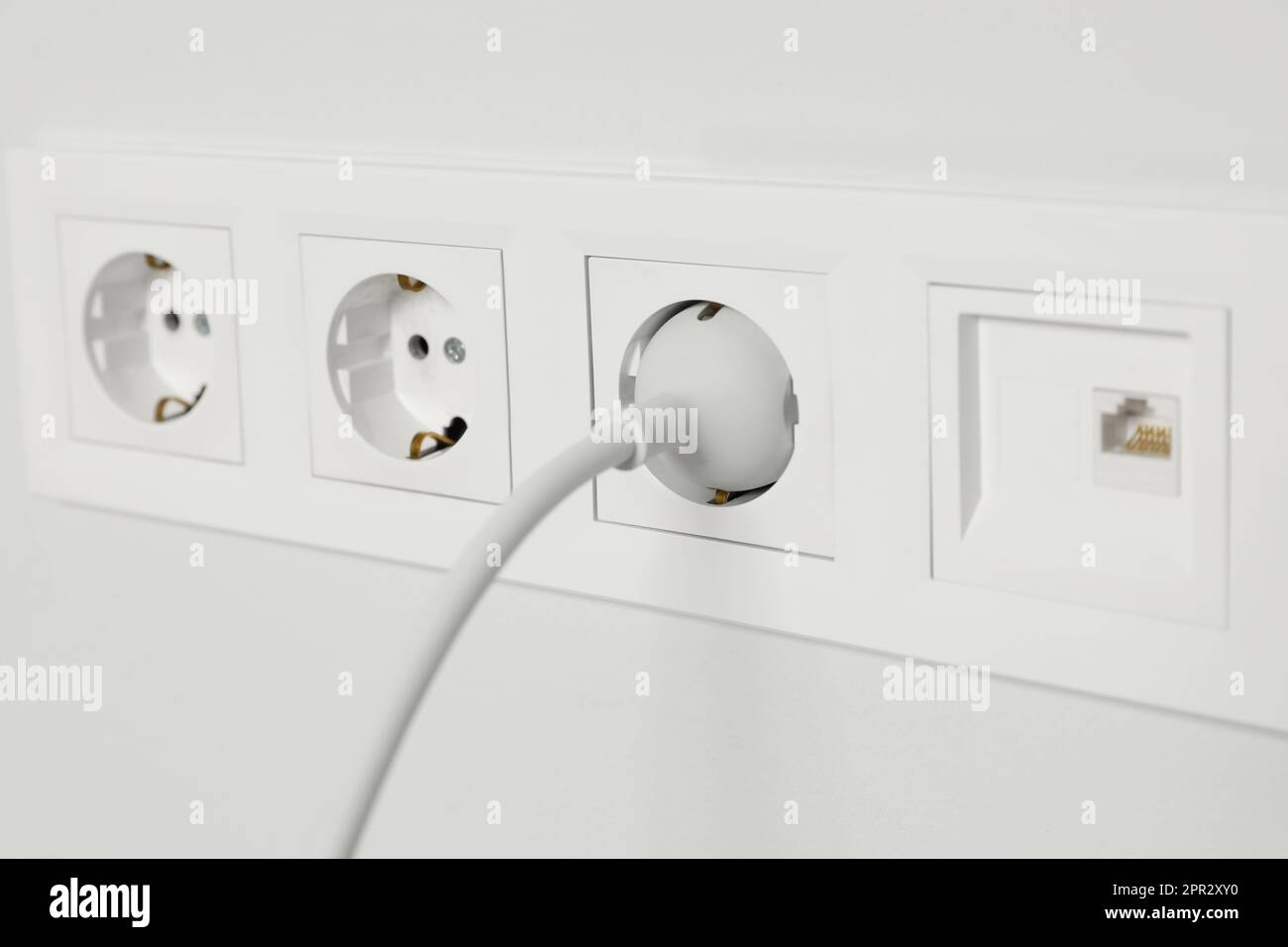 Power sockets with inserted plug on white wall, closeup. Electrical