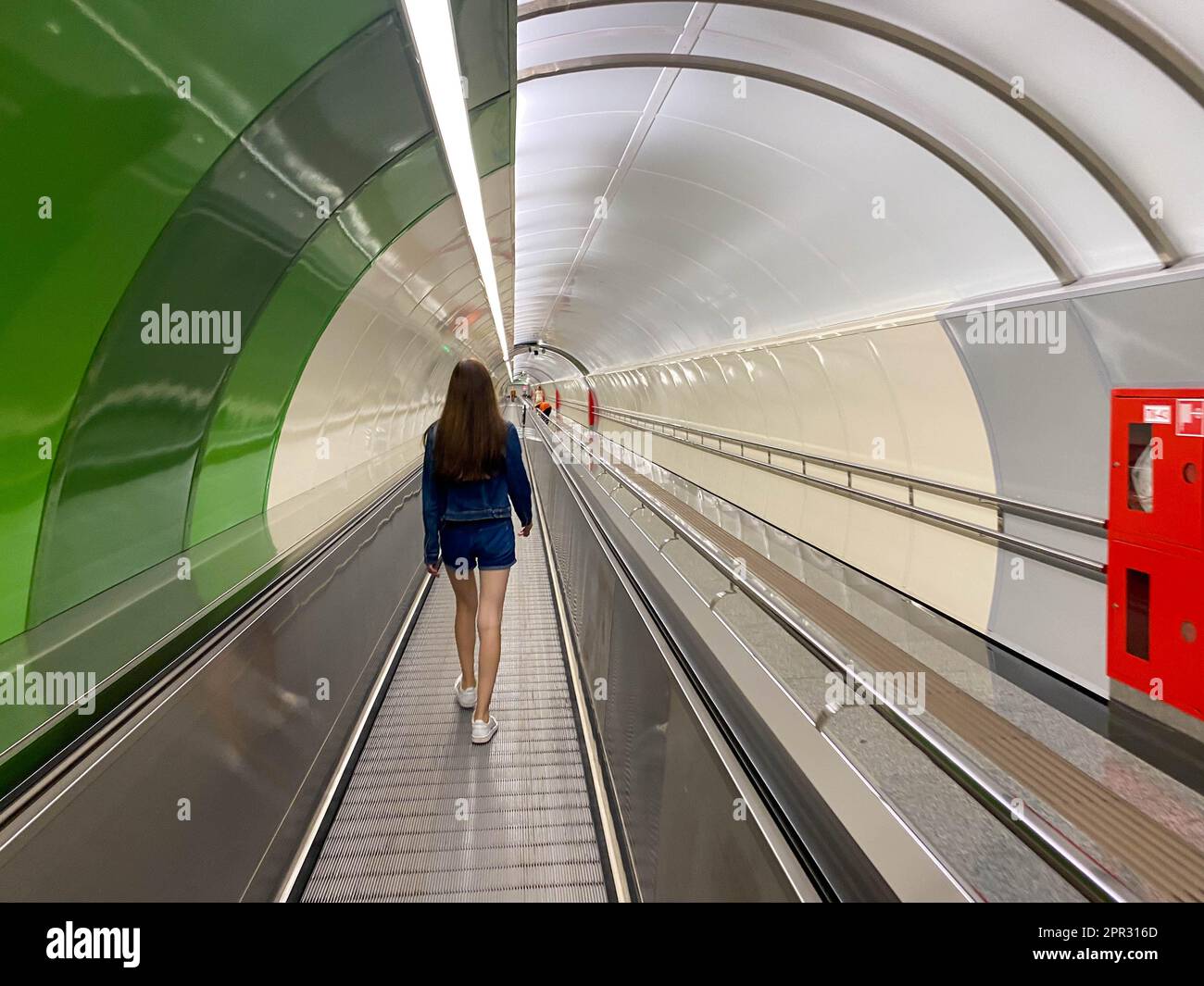 A beautiful girl walks along the travolator in the subway tunnel to go to another underground station. View from the back. Stock Photo