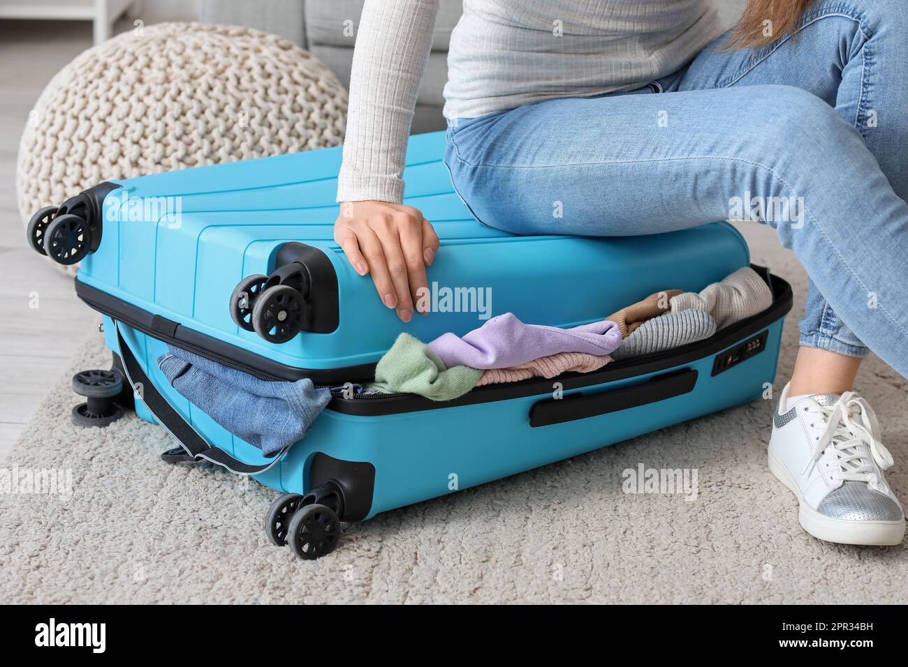 Young woman trying to close suitcase with a lot of clothes at home