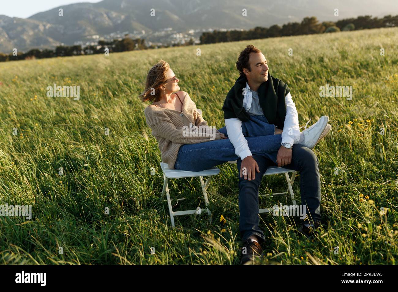 Young happy couple sitting on chairs in feather grass field on summer sunset day. Stock Photo