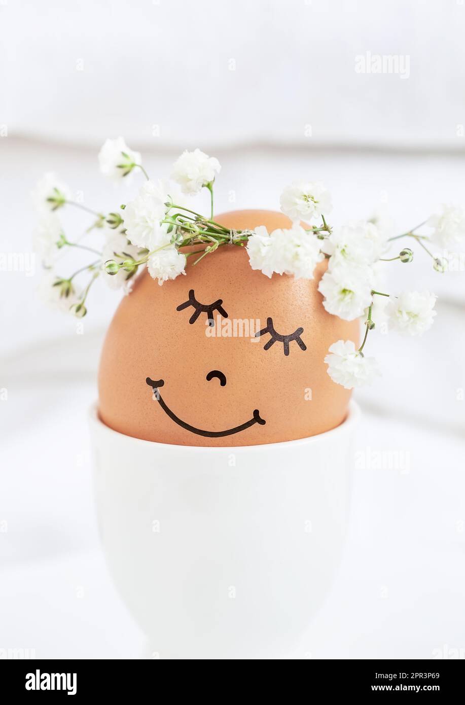 Easter egg with cute face in floral wreath crowns in egg cup on white background. Easter egg with flowers and sleepy eyes in sunny light. Happy Easter Stock Photo