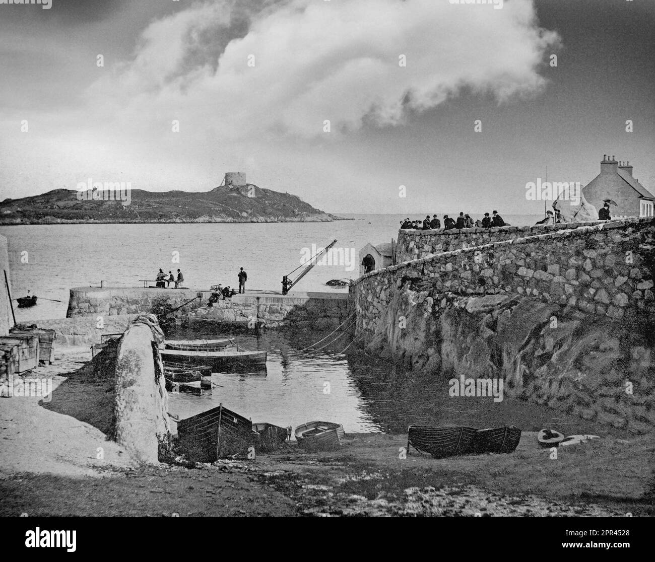A late 19th century photograph of Coliemore Harbour, also known historically as Dalkey Harbour in County Dublin, Ireland. The harbour developed strategic value as a port for bulk shipments bound for Dublin during the 15th century. The channel between Dalkey Island with its Martello Tower and the mainland provided ideal conditions for unloading galleons carrying heavy cargo due to its depth (relative to Dublin Bay) and its sheltered position. The treacherous shallows of Dublin Bay prevented direct shipments into the city center, making Dalkey an ideal access point for trade. Stock Photo