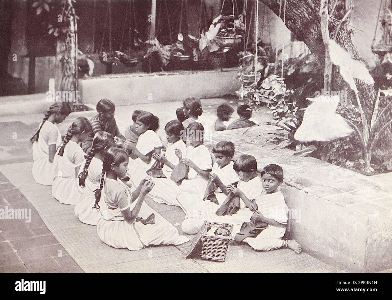 A sewing class in the courtyard at the Dohnavur compound. Half-tone engraving from a photograph by Mr. Penn of Ootacamund (now known as Ooto), in the Tamil Nadu region of Southern India, c1912. The image relates to the Church of England Zenana Mission’s nursery and compound in Dohnavur, also in Tamil Nadu and about thirty miles from the southern tip of India. The compound was run by Amy Wilson-Carmichael and sought to promote Christianity among babies and children (at the time only girls, many who were victims of temple prostitution). Stock Photo