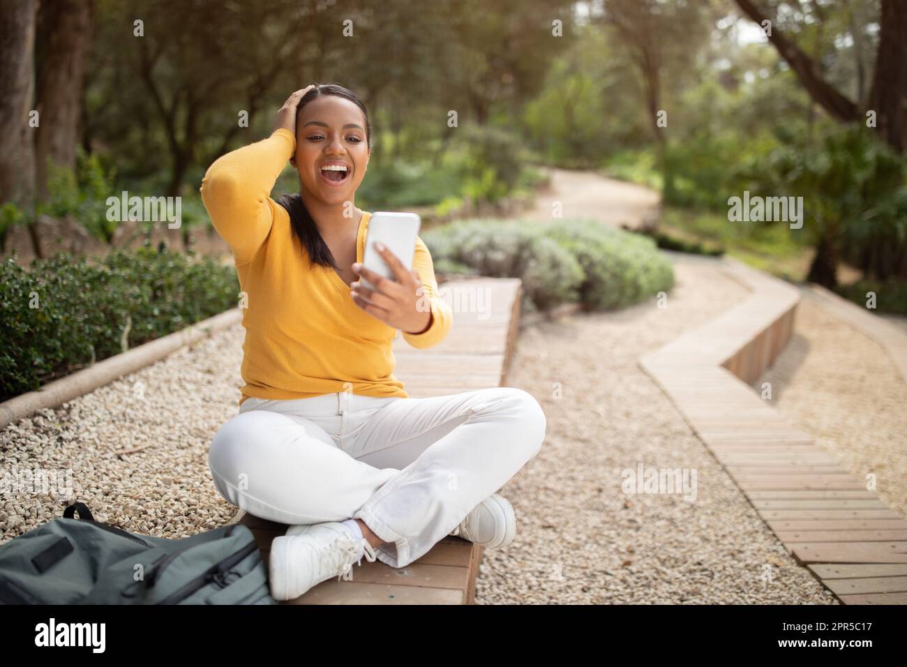 Wow. Excited young brazilian lady reading great news on smartphone and touching head, sitting in park outdoors Stock Photo