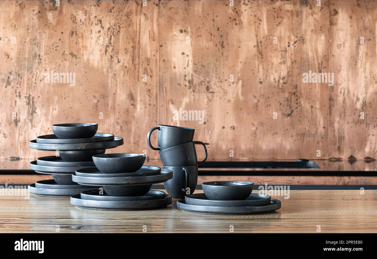 A stack of rustic black ceramic plates, cups and bowls on a wooden table in a modern kitchen. Stock Photo