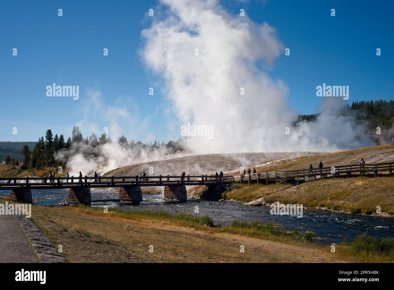 The Excelsior Geyser erupts dramatically above a footbridge across the Firehole River in Yellowstone National Park. Stock Photo