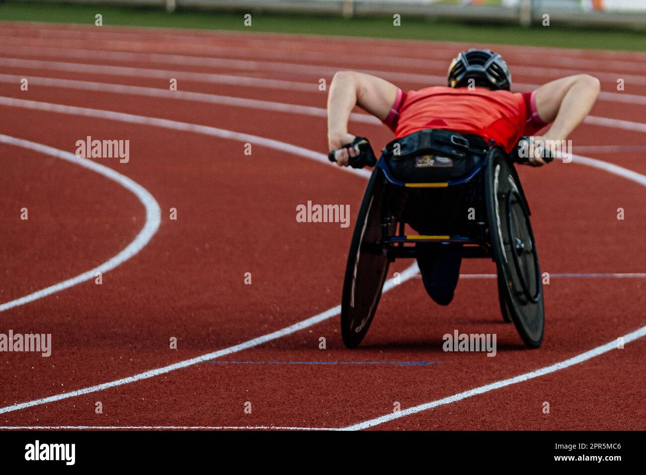 rear view male athlete in racing wheelchair riding on red track stadium, summer para athletics championships Stock Photo