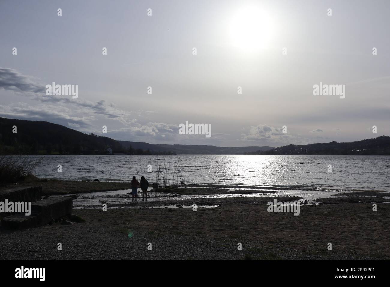 White April sunset at the lower Lake Constance. In front of it the small silhouettes of two children playing in the silvery Feldbach estuary delta. Stock Photo