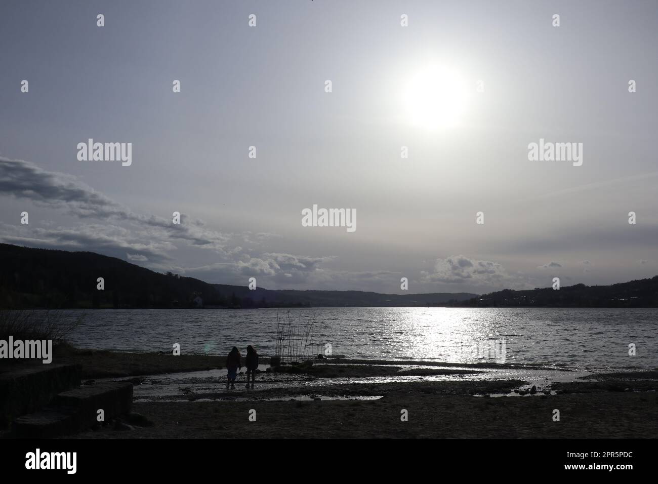 White April sunset at the lower Lake Constance. In front of it the small silhouettes of two children playing in the silvery Feldbach estuary delta. Stock Photo