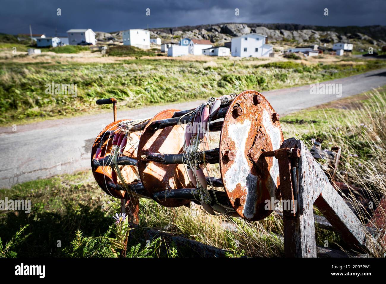 Rustic hand reel from a commercial fishing boat lying in tall grass overlooking small wooden homes at Keels Newfoundland Canada along the Discovery Tr Stock Photo
