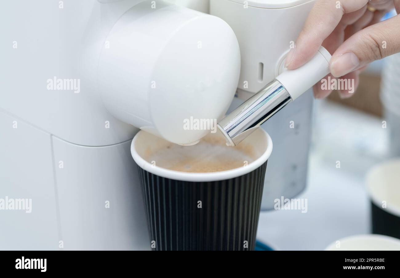 Woman making a cup of hot coffee with capsule coffee machine. Woman hand holding frothed milk dispenser of capsule coffee machine on table. Espresso coffee maker. Morning drink. Modern home equipment. Stock Photo