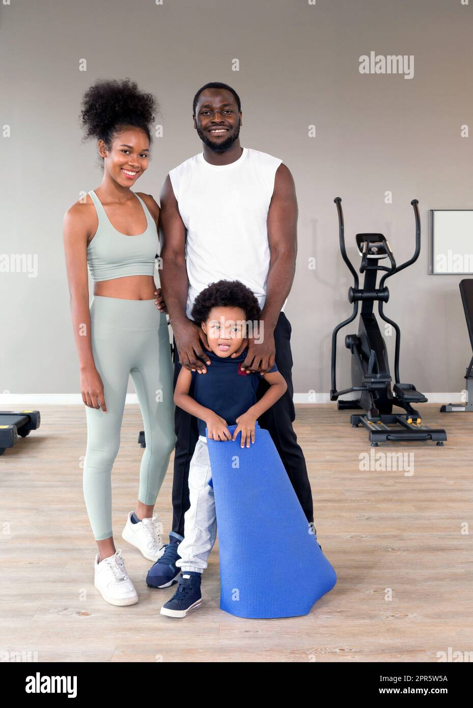 Happy family enjoy holiday together in fitness center. Young father hold his son shoulder while the boy try to play with yoga mat. Stock Photo