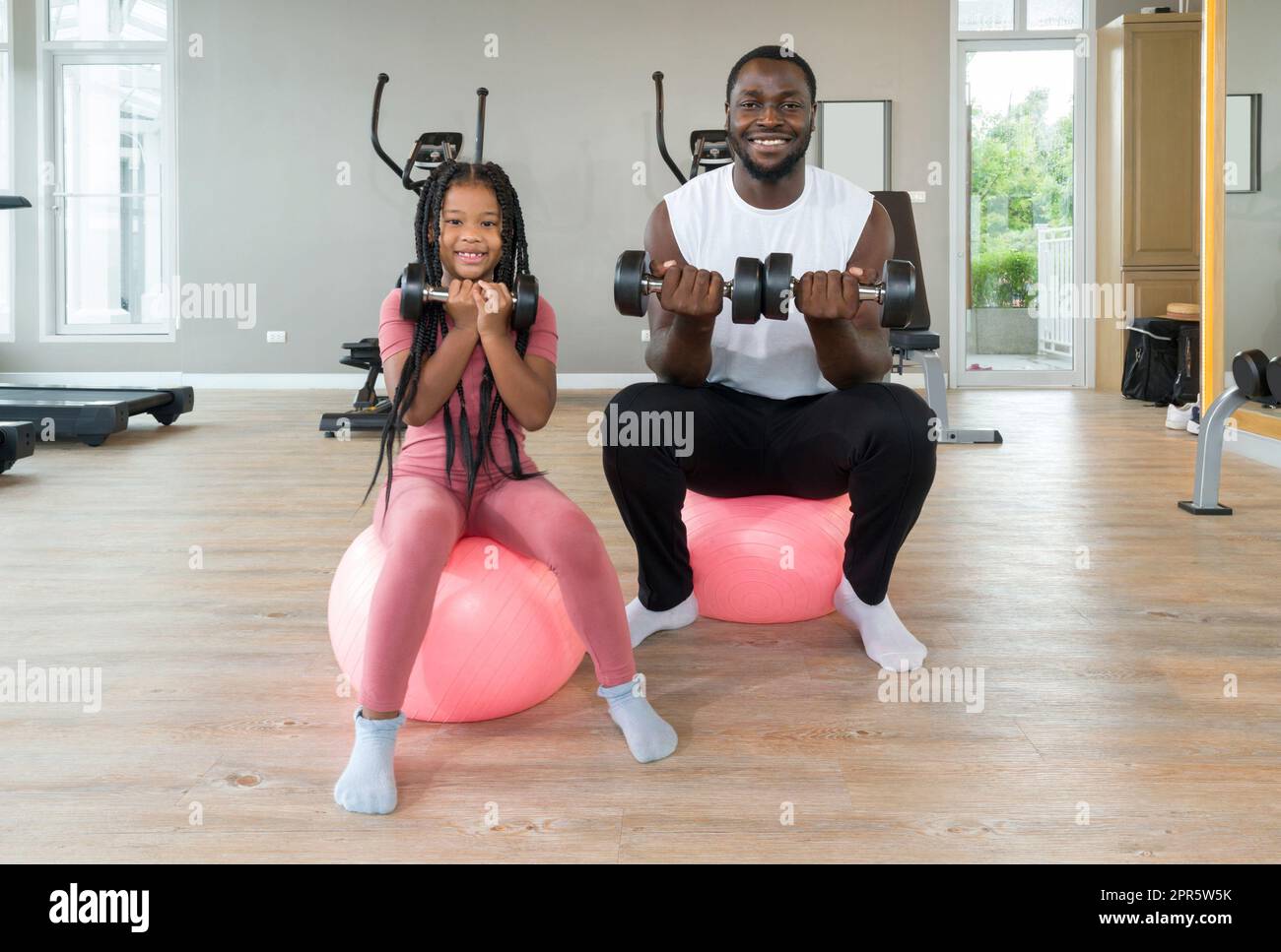 Young short curly black hair man and his daughter lifting barbell with both hands while sitting on yoga ball. Happy family enjoy holiday together in fitness center. Stock Photo