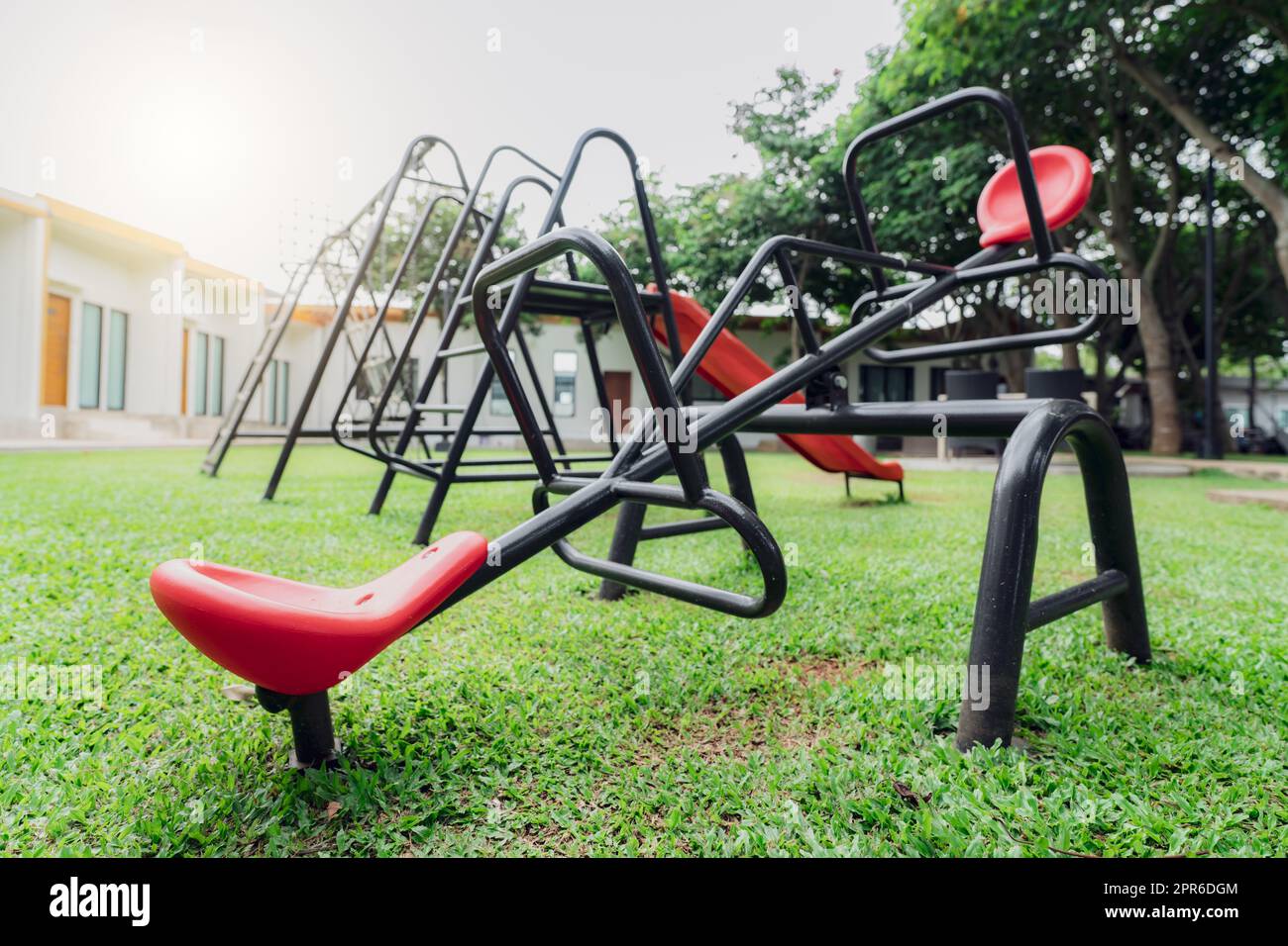 Red seesaw in the playground. Playground equipment for children to play. Plastic seesaw or teeter-totter, swing, and slide at outdoor playground with green grass ground. Outdoor kids toy at park. Stock Photo