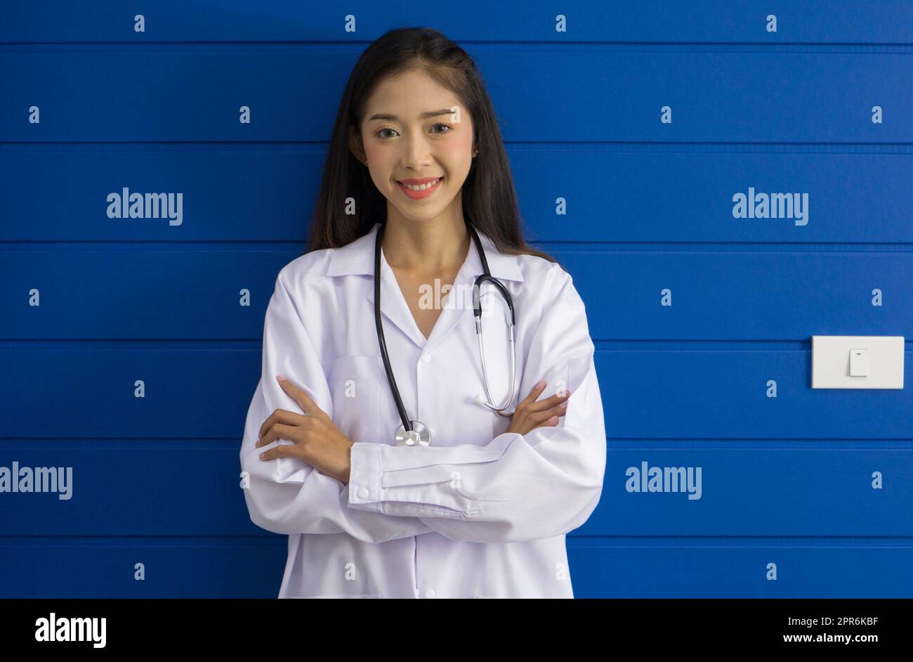 Young asian doctor in white gown and stethoscope stand with arm cross in front of blue wall. Stock Photo